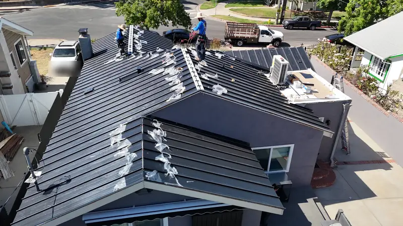 Roofers installing black standing seam metal panels with protective film still on, over a gray stucco house